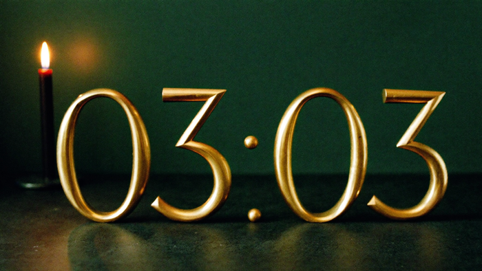 Messingziffern 03:03 auf einem dunklen Steinsims vor einer dunkelgrünen Wand, links eine brennende Kerze – Titelbild zur Spiegelstunde 03:03 Uhr