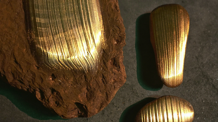 Brauner Bronzit-Rohstein mit faserig schimmernder Bronze-Oberfläche und zwei polierte Bronzit-Steine auf dunklem Stein im Kerzenlicht