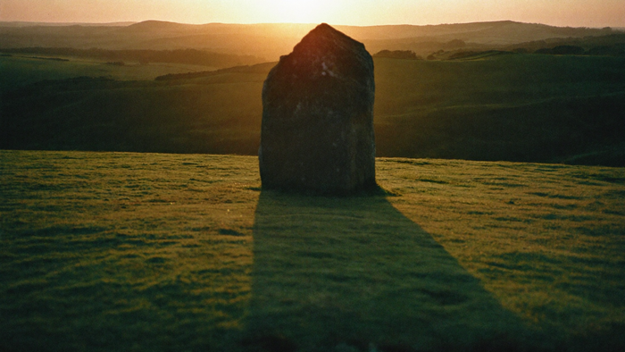 Verwitterter Menhir auf einem Grasrücken im Morgenlicht, ein langer gerader Schatten läuft über die Wiese