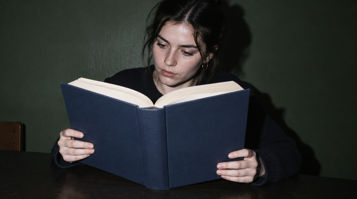 Eine junge Frau liest konzentriert in einem sehr dicken, aufgeschlagenen Buch mit schlichtem dunkelblauem Einband vor dunkelgrüner Wand