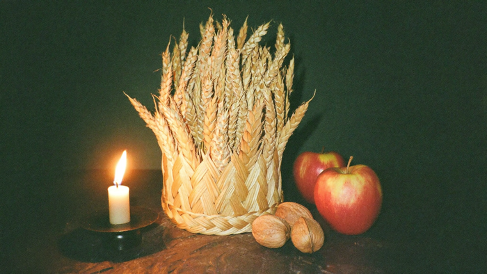 Geflochtene Erntekrone aus Weizenähren neben Kerze, Äpfeln und Walnüssen auf dunklem Holz