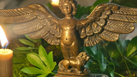 Bronzene Engelsfigur mit ausgebreiteten Flügeln, zu ihren Füßen ein kleiner Löwe, zwischen grünen Blättern im Kerzenlicht – Titelbild zu Erzengel Ariel, dem „Löwen Gottes“