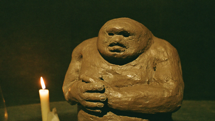 Grob geformte Lehmfigur eines Golems mit schwerem Kopf, hohlen Augen und verschränkten Händen, links eine brennende Kerze vor dunkelgrüner Wand