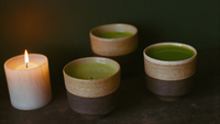 Drei handgedrehte Keramiktassen mit Matcha auf dunklem Stein, links eine brennende Kerze vor dunkelgrüner Wand