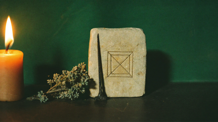 Weathered stone talisman with an incised geometric square, an iron nail and dried rue beside a warm candle against a dark green wall