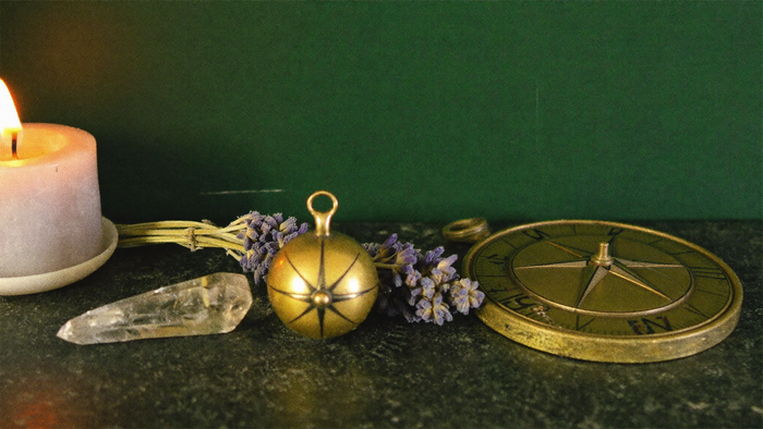 Messing-Armillarsphäre, ein antiker Kompass, ein klarer Bergkristall und getrockneter Lavendel auf dunkler Steinfläche neben einer warmen Kerze vor dunkelgrüner Wand