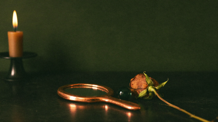 Kupferner Handspiegel neben einer getrockneten Rose und einer grünen Glasperle im Kerzenlicht
