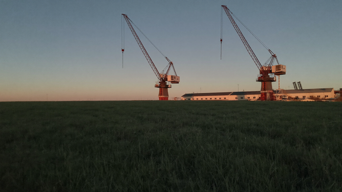 Weite Steppe zur blauen Stunde, am Horizont zwei hohe Portalkräne und flache Industriegebäude im letzten Licht