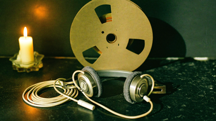 Vintage-Kopfhörer aus den 1970ern und eine Tonbandspule auf dunklem Stein, links eine brennende Kerze, dahinter eine dunkelgrüne Wand im Schatten