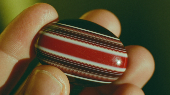Nahaufnahme eines polierten Sardonyx-Cabochons mit geraden roten, braunen und weißen Bändern zwischen zwei Fingerspitzen bei Kerzenlicht