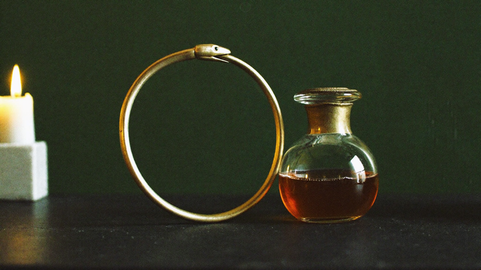Bronzener Ouroboros-Ring, eine Schlange, die sich in den Schwanz beißt, neben einer Glasflasche mit bernsteinfarbener Flüssigkeit im Kerzenlicht