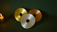 Drei Metallscheiben in Gold, Silber und Kupfer im Kerzenlicht vor grüner Wand