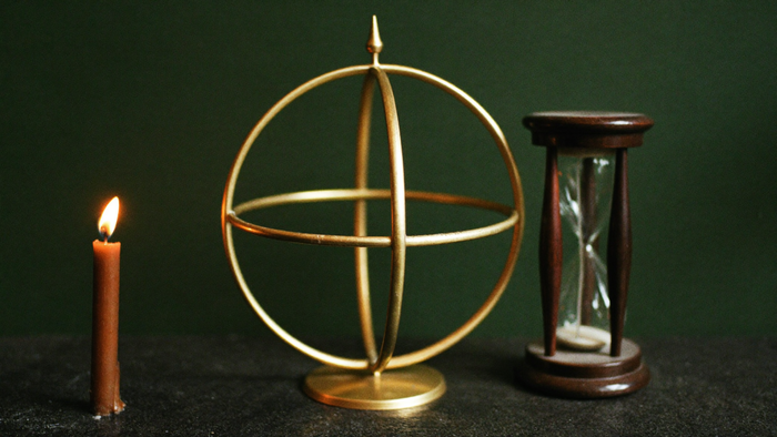 Eine schlichte Armillarsphäre aus Messing steht neben einer kleinen Sanduhr auf dunklem Stein, links brennt eine Kerze vor einer dunkelgrünen Wand.
