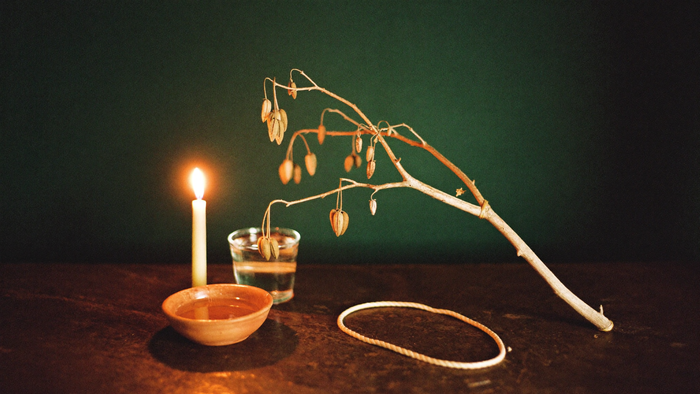 Eschenzweig mit Samen, eine Tonschale mit Wasser und ein Seil im Kerzenlicht vor dunkelgrüner Wand