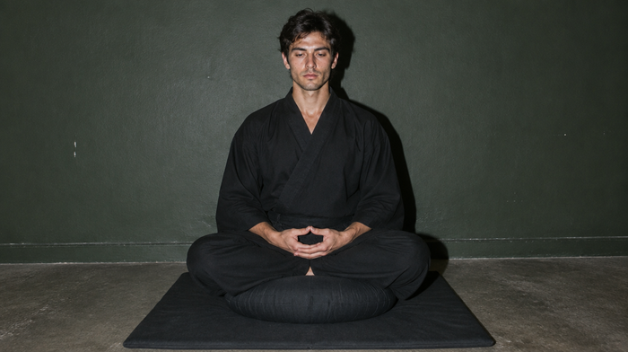 Zazen: junger Mann in schwarzer Robe sitzt aufrecht auf einem runden Zafu-Kissen, die Hände im kosmischen Mudra, vor einer dunkelgrünen Wand