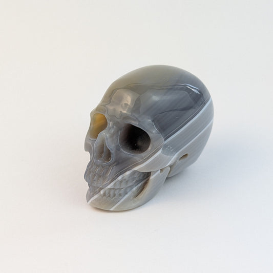 Ein 84g schwerer, handgeschnitzter Kristallschädel 'Ianna' aus Botswana Achat mit feinen, sanften Bändern in Grau- und Rosatönen.