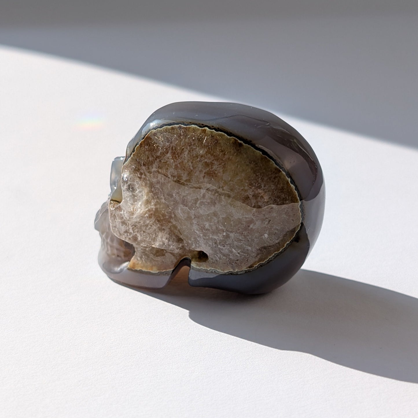 Ein 86g schwerer Kristallschädel 'Der Wächter der zwei Welten' aus Gebändertem Achat, dessen eine Hälfte eine offene, funkelnde Druzy-Geode ist.