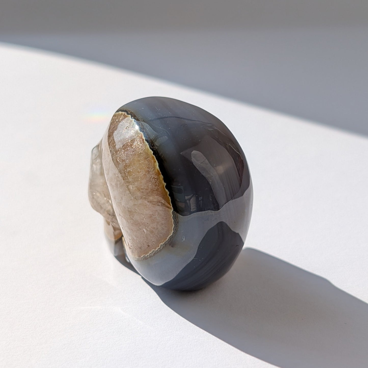 Ein 86g schwerer Kristallschädel 'Der Wächter der zwei Welten' aus Gebändertem Achat, dessen eine Hälfte eine offene, funkelnde Druzy-Geode ist.