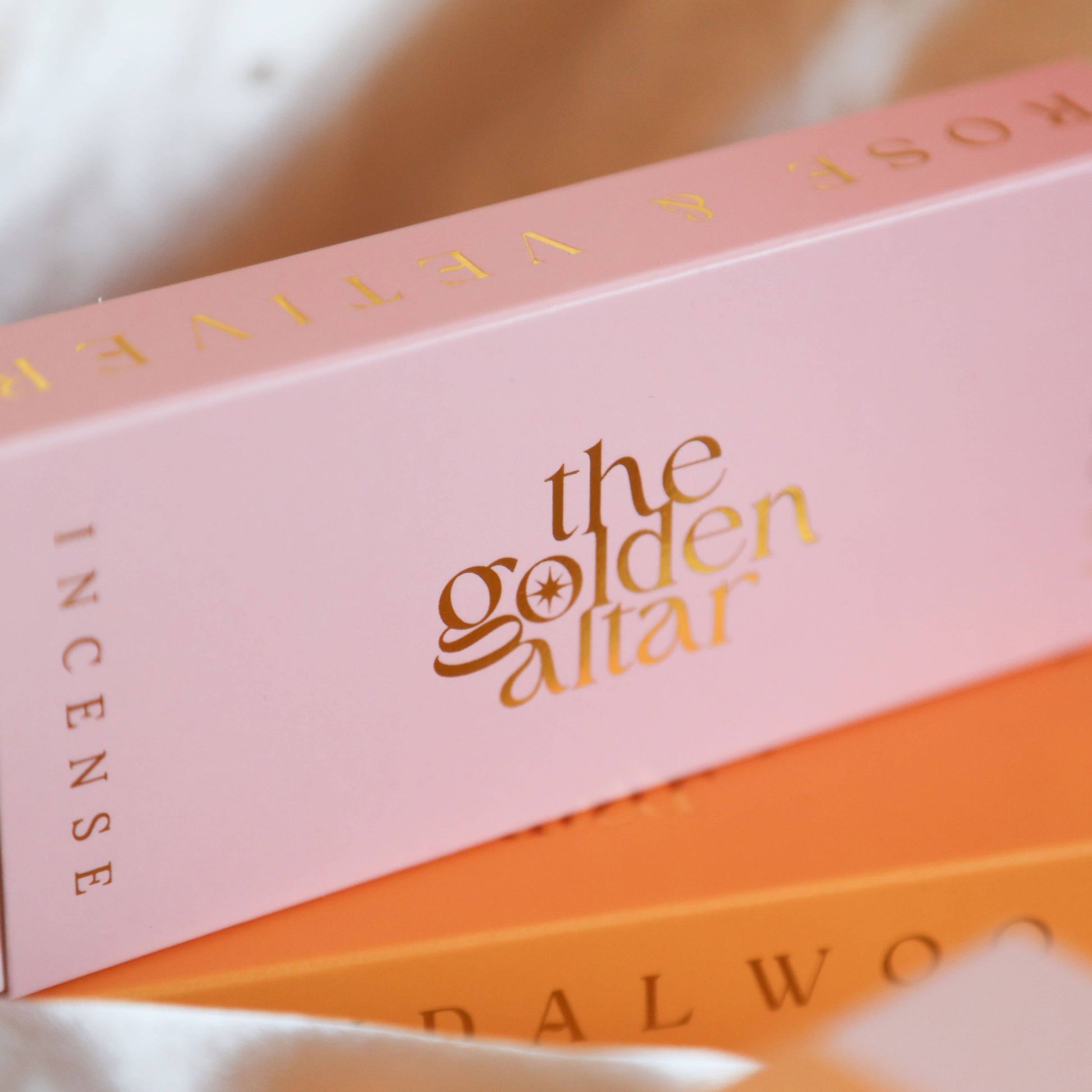 Eine rosa-orangefarbene Box von The Golden Altar mit 30 Rose & Vetiver Räucherstäbchen, die daneben liegen.