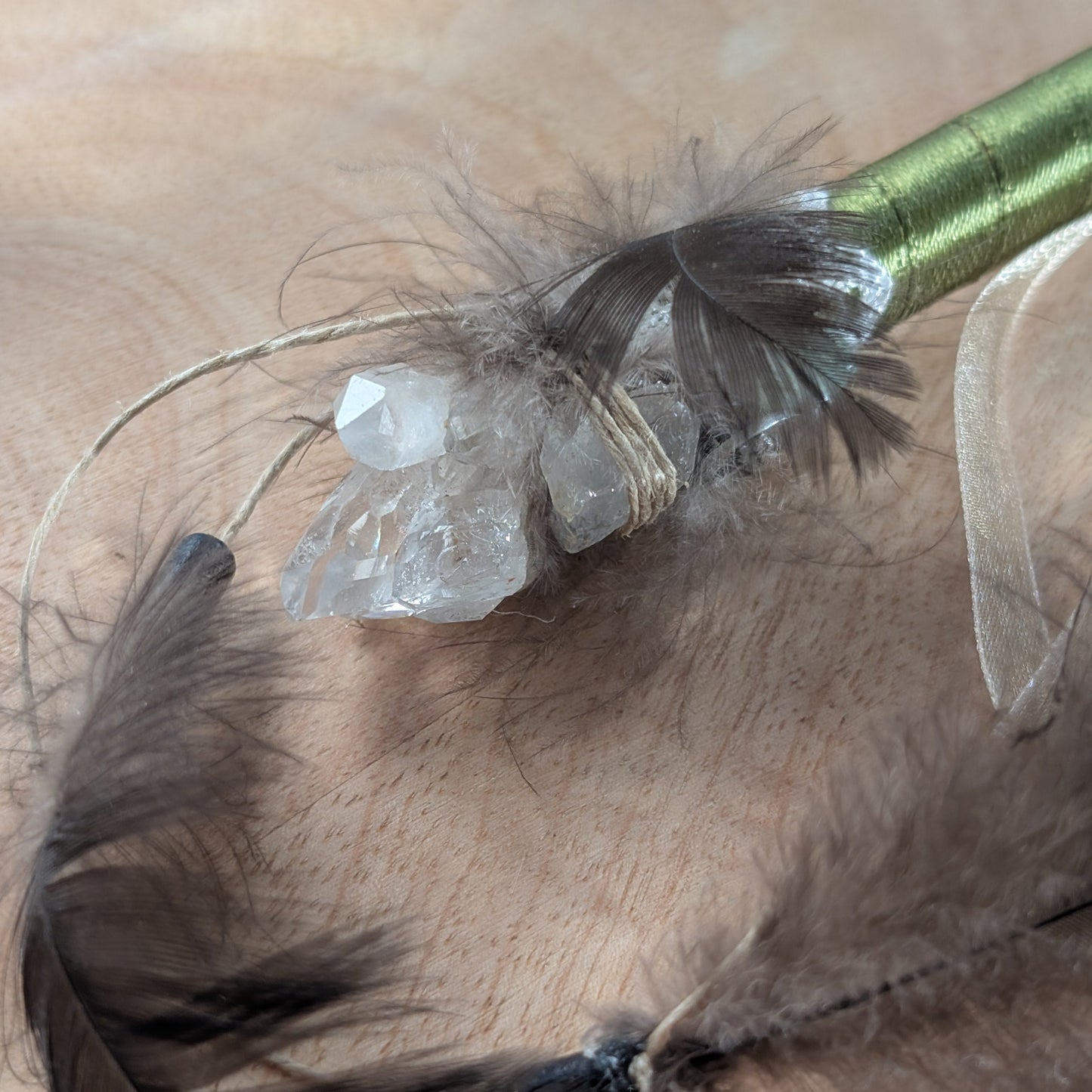 Handgefertigter Zauberstab mit einem goldgelben Citrin und schimmerndem Drusenquarz, verziert mit Federn und einem grünen Band, auf einer Holzplatte.