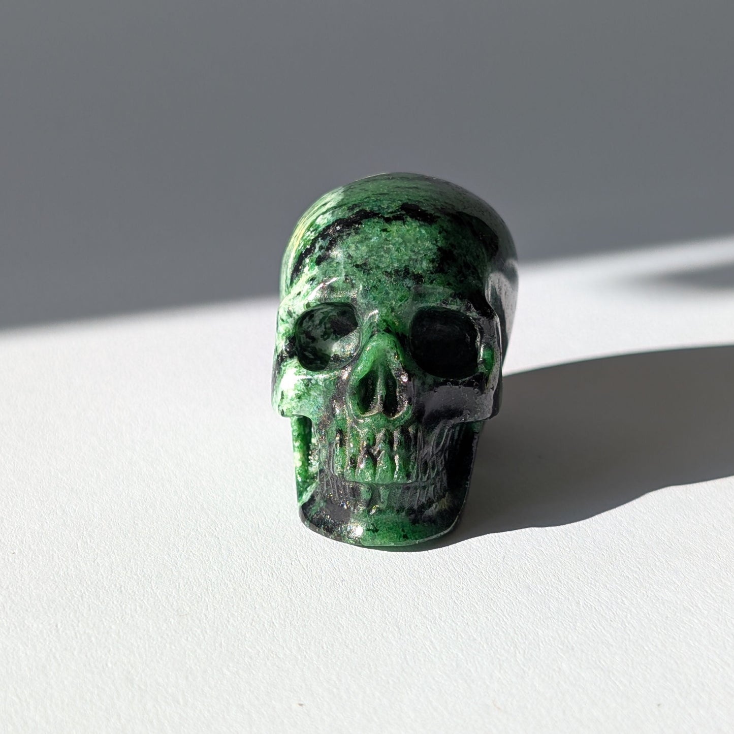 Kleiner Kristallschädel "Leonidar" (92g) aus grünem Zoisit mit schwarzen Einschlüssen.