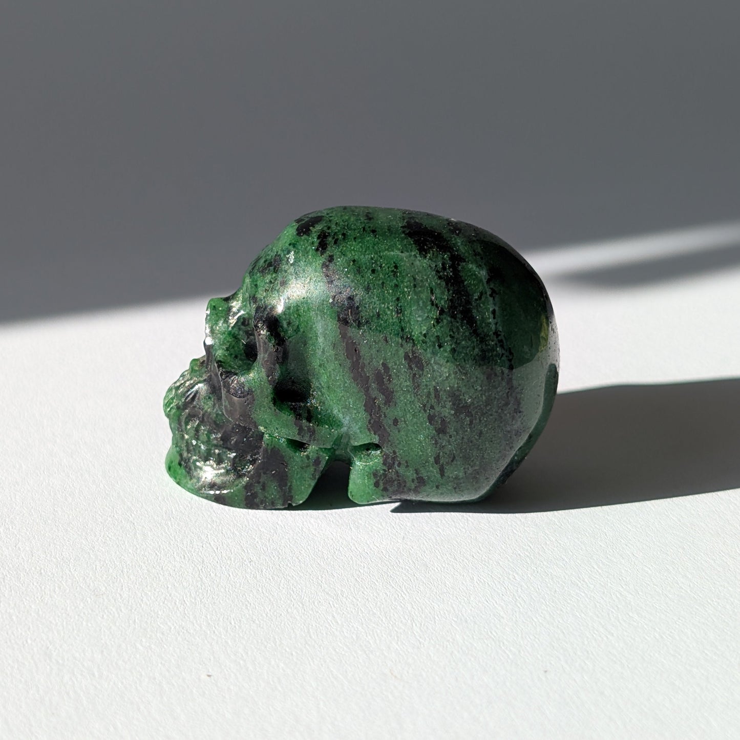 Kleiner Kristallschädel "Leonidar" (92g) aus grünem Zoisit mit schwarzen Einschlüssen.
