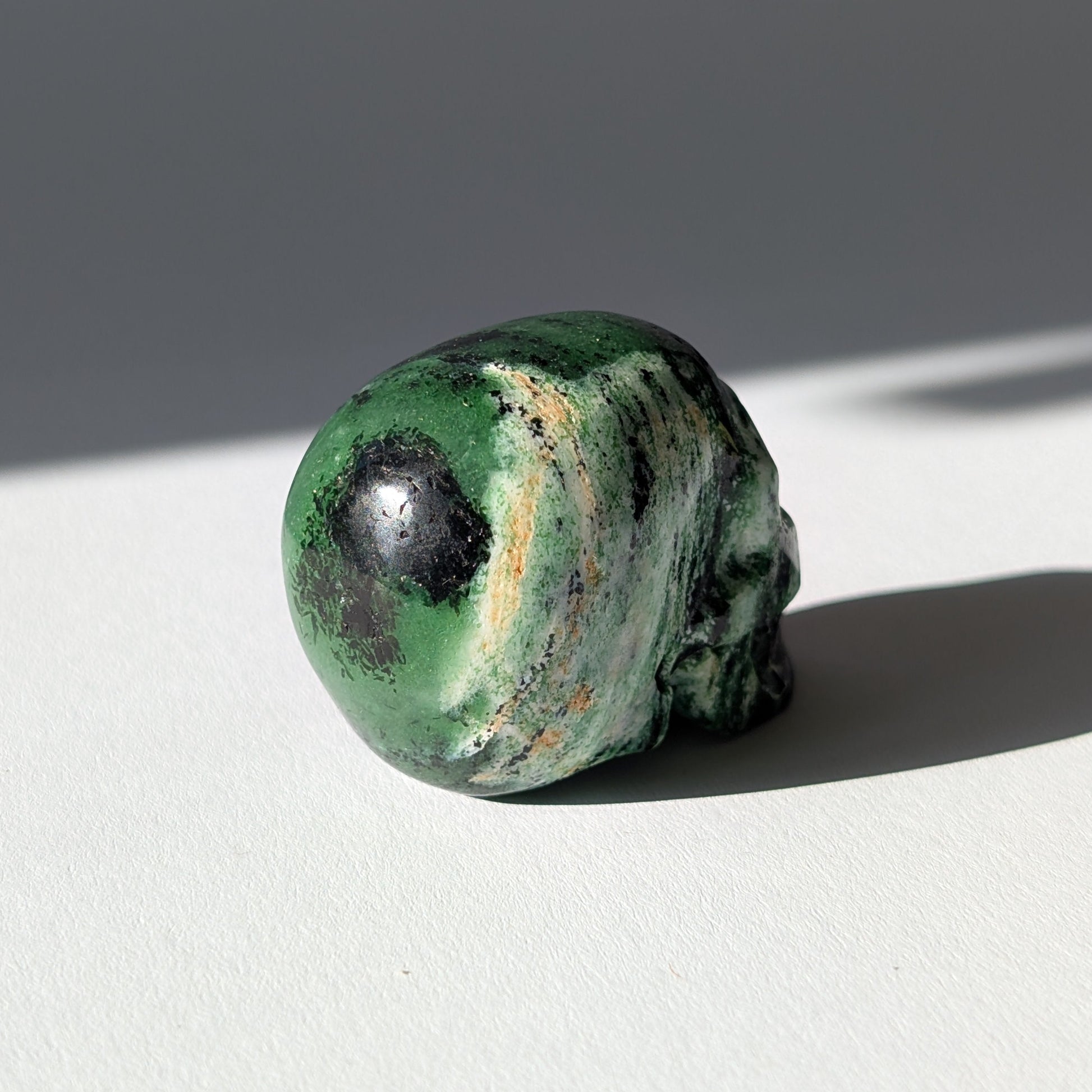 Kleiner Kristallschädel "Leonidar" (92g) aus grünem Zoisit mit schwarzen Einschlüssen.