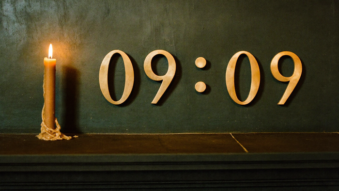 Messingziffern 09:09 an einer dunkelgrünen Wand über einem Steinsims, links eine brennende Bienenwachskerze – Titelbild zur Spiegelstunde 09:09 Uhr