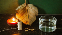 Getrocknetes Eichenblatt, eine Rolle ungebleichter Leinenfaden, ein alter Messingschlüssel und eine blanke Glasschale mit Wasser auf dunklem Stein, links eine brennende Kerze vor dunkelgrüner Wand
