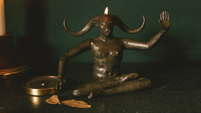 Bronzefigur des Baphomet mit Bockskopf, gedrehten Hörnern, Flamme zwischen den Hörnern und Pentagramm auf der Stirn, eine Hand erhoben, eine gesenkt — bei Kerzenlicht vor dunkelgrüner Wand