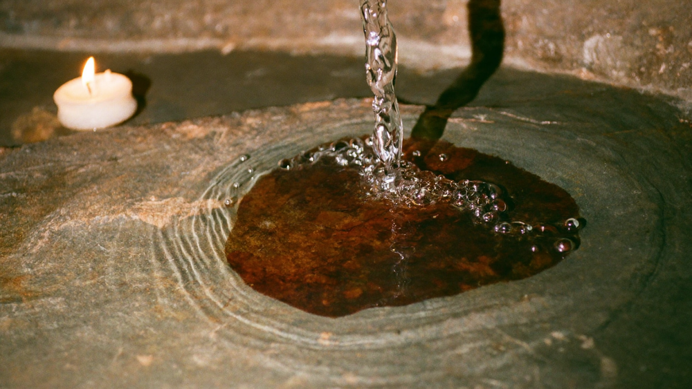 Klares Wasser läuft über rostrot verfärbten Stein in eine flache Steinschale, von einer Kerze beleuchtet