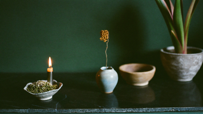 Kleine Vase mit Trockenblume, Keramikschale, brennende Kerze und Übertopf auf einer dunklen Steinfläche als Hausaltar