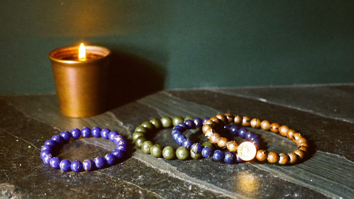Vier Edelstein-Armbänder in Violett, Blau, Grün und Goldbraun auf dunklem Stein neben einer brennenden Kerze