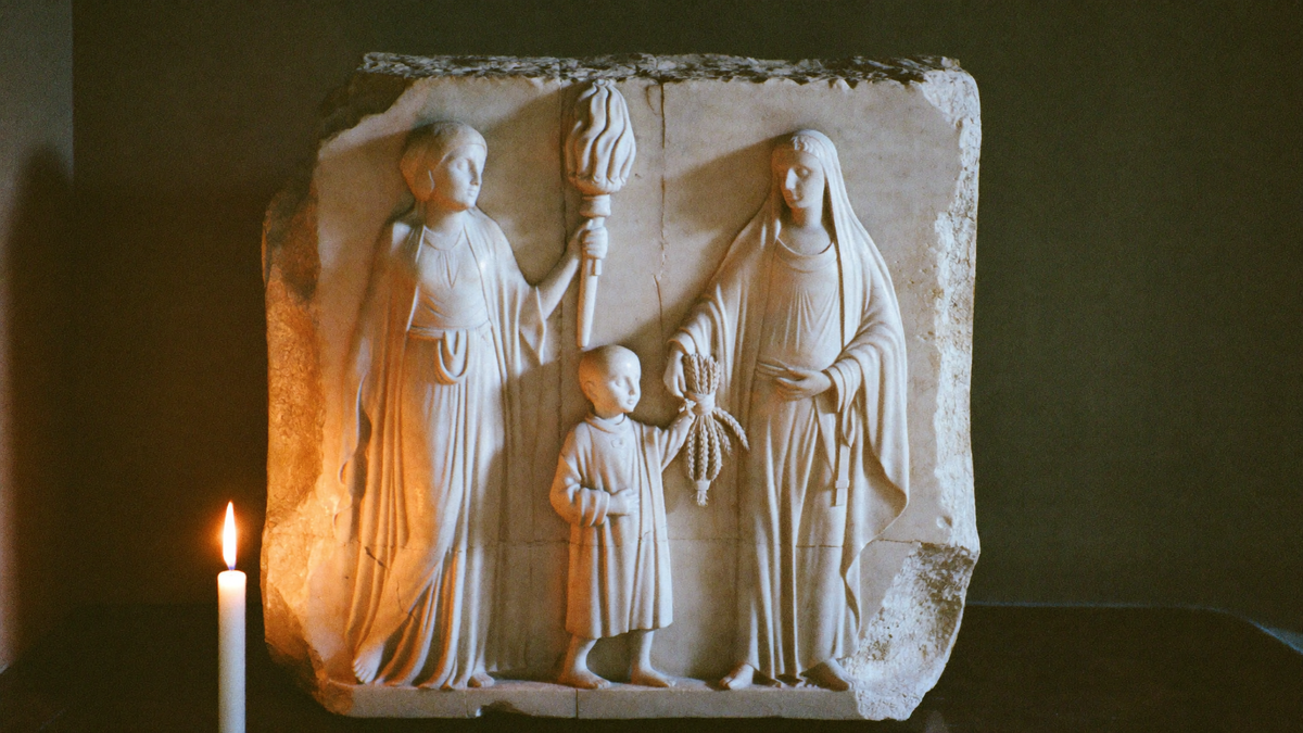 Marmorrelief im Kerzenlicht: eine Göttin mit Fackel, eine zweite reicht einem Knaben ein Bündel Ähren — Demeter, Persephone und Triptolemos