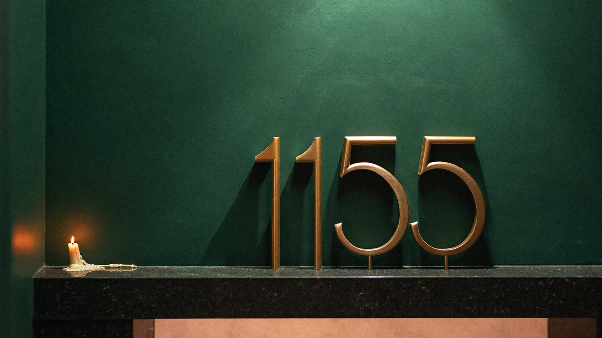 Messingziffern 1155 an einer dunkelgrünen Wand über einem Steinsims, links eine brennende Kerze – Titelbild zur Engelszahl 1155