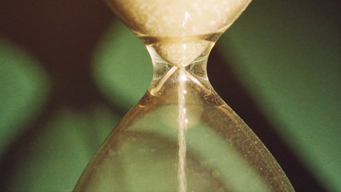 Nahaufnahme einer Sanduhr, durch deren Hals die letzten Sandkörner rieseln, im Kerzenlicht vor dunkelgrünem Hintergrund – Titelbild zur Engelszahl 9999