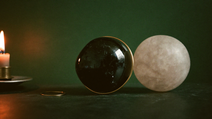 Eine schwarze polierte Obsidiankugel und eine helle Mondsteinkugel nebeneinander auf dunklem Stein, davor ein dünner Messingring, links eine brennende Kerze vor dunkelgrüner Wand