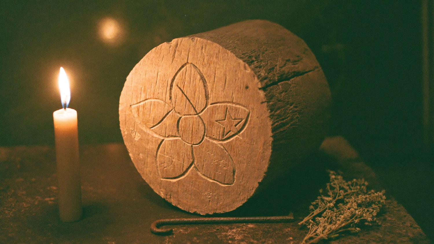 In einen alten Holzbalken geritzte Sechspass-Rosette mit fünfzackigem Stern, daneben eine brennende Kerze, ein Eisennagel und getrocknete Kräuter