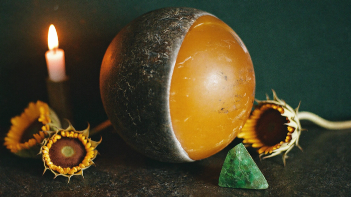 Goldene Kugel halb verdeckt von einem dunklen Stein als Sinnbild der Sonnenfinsternis, daneben getrocknete Sonnenblumen, ein grüner Kristall und eine brennende Kerze