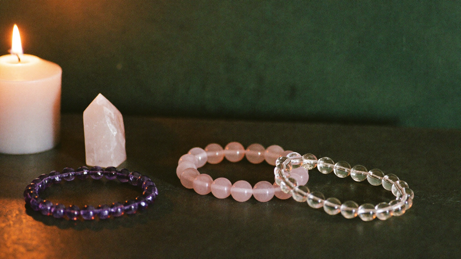 Drei Edelstein-Armbänder aus Amethyst, Rosenquarz und Bergkristall bei Kerzenlicht vor dunkelgrüner Wand
