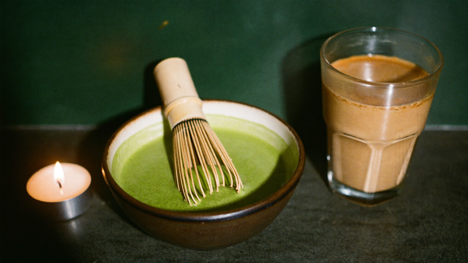 Schale mit grünem Matcha und Bambusbesen neben einem Glas Chai Latte bei Kerzenlicht vor dunkelgrüner Wand