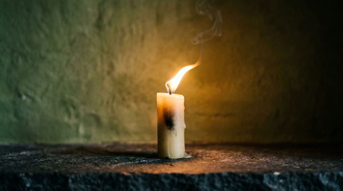 Ritualkerze mit gebogener Flamme und aufsteigendem Rauchfaden auf dunklem Stein vor dunkelgrüner Wand