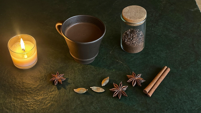 Tasse heißer Kakao, ein Glas gemahlene Gewürze, Sternanis, Kardamom und Zimtstange neben einer brennenden Kerze auf dunklem Stein