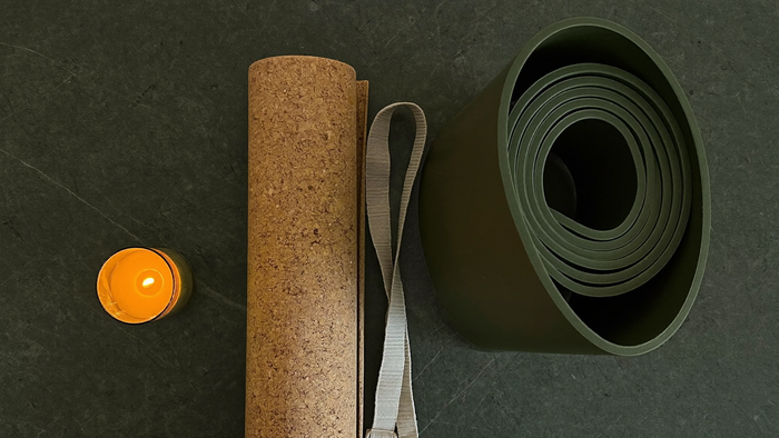 Eingerollte Kork-Yogamatte und dunkelgrüne Naturkautschukmatte neben einer Kerze auf dunklem Stein