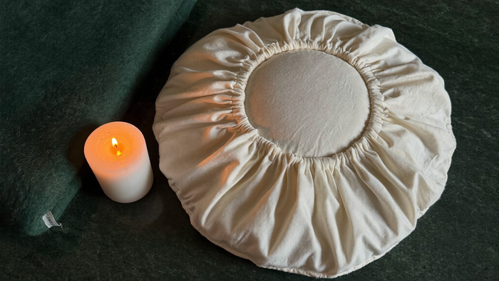 Rundes Zafu-Meditationskissen aus Baumwolle neben einer brennenden Kerze und einer grünen Wolldecke auf dunklem Untergrund