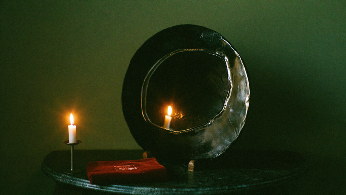 Ein runder schwarzer Obsidianspiegel steht aufrecht auf einem dunklen Steintisch und spiegelt eine Kerzenflamme, davor ein rotes Samttuch, dahinter eine dunkelgrüne Wand