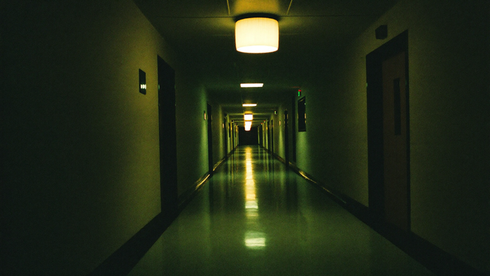 Leerer Klinikflur bei Nacht, eine einzelne warme Deckenleuchte am Ende des Ganges, der Rest liegt in grünem Schatten