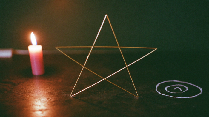 Aufrechtes Pentagramm aus Messingdraht auf dunklem Stein, daneben ein Kreidekreis, links eine brennende Kerze vor dunkelgrüner Wand