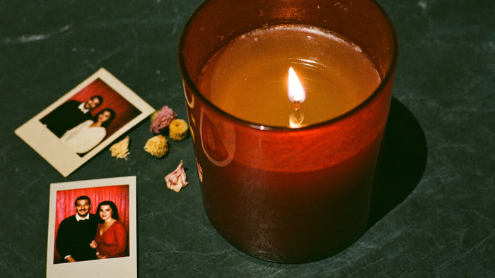 Brennende Kerze in rubinrotem Glas neben zwei verblassten Familienfotos und getrockneten Chrysanthemenblüten auf dunklem Stein