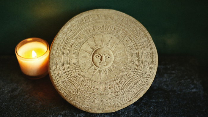 Runder Maya-Kalenderstein mit Tageszeichen und Sonnengesicht neben einer brennenden Kerze – Portaltage Juli 2027